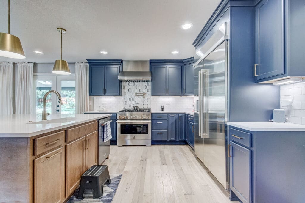 Modern kitchen remodel with bold blue cabinetry and stainless steel appliances – This FBC Remodel project features a striking combination of deep blue cabinets, a sleek white island with wood accents, and a professional-grade stainless steel range and refrigerator. The custom kitchen design blends modern elegance with functional space, perfect for any home chef. Gold pendant lighting and hardware add a luxurious touch, while the light wood flooring provides warmth and balance to the space. Ideal for anyone looking for a high-end, stylish kitchen renovation.
