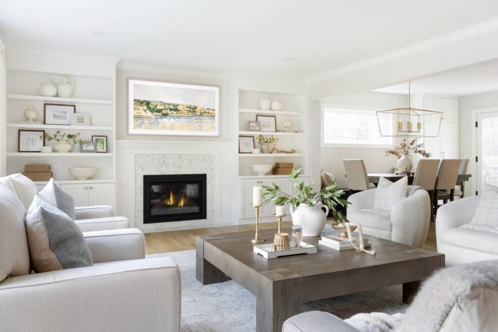 A bright, open living room with a cozy fireplace, custom white built-in shelves, and neutral-toned furniture arranged around a wood coffee table with candles and greenery.