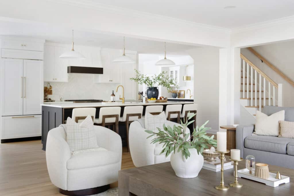 Open-concept living space featuring plush swivel chairs, a rustic coffee table with decor, and a view into a modern kitchen with white cabinetry and gold accents.