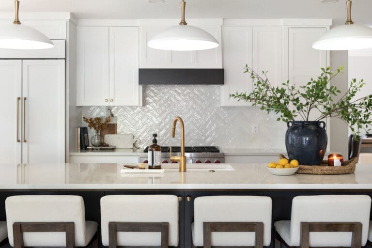 A bright kitchen island with a gold faucet, bowl of lemons, candle, and a large ceramic vase with branches, set against a white herringbone tile backsplash. broomfield kitchen remodeling