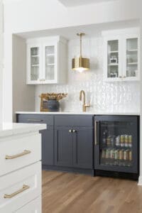 Chic beverage bar with navy lower cabinets, white upper cabinets with glass doors, gold hardware, and a beverage fridge stocked with canned drinks.