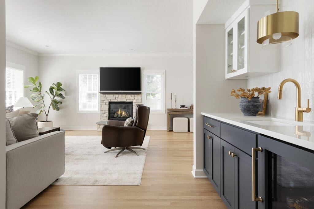 A cozy lounge space with a leather armchair facing a stone fireplace and TV, adjacent to a stylish beverage bar with gold fixtures and navy cabinetry.