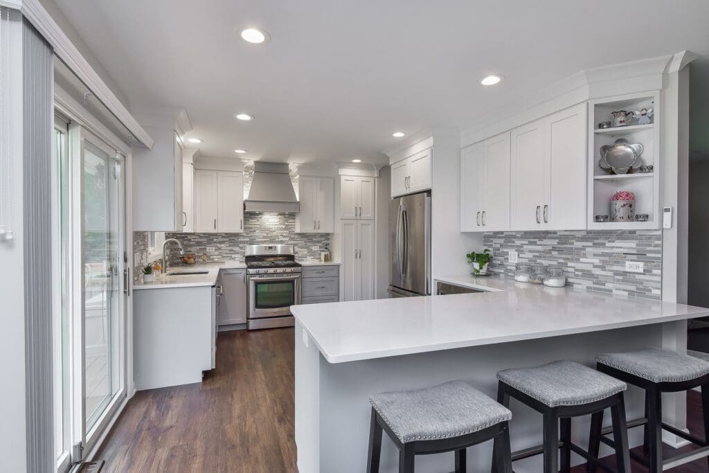 A modern open-concept kitchen with white shaker cabinets, stainless steel appliances, quartz countertops, and a glass tile backsplash. A large island with gray upholstered stools offers casual seating.