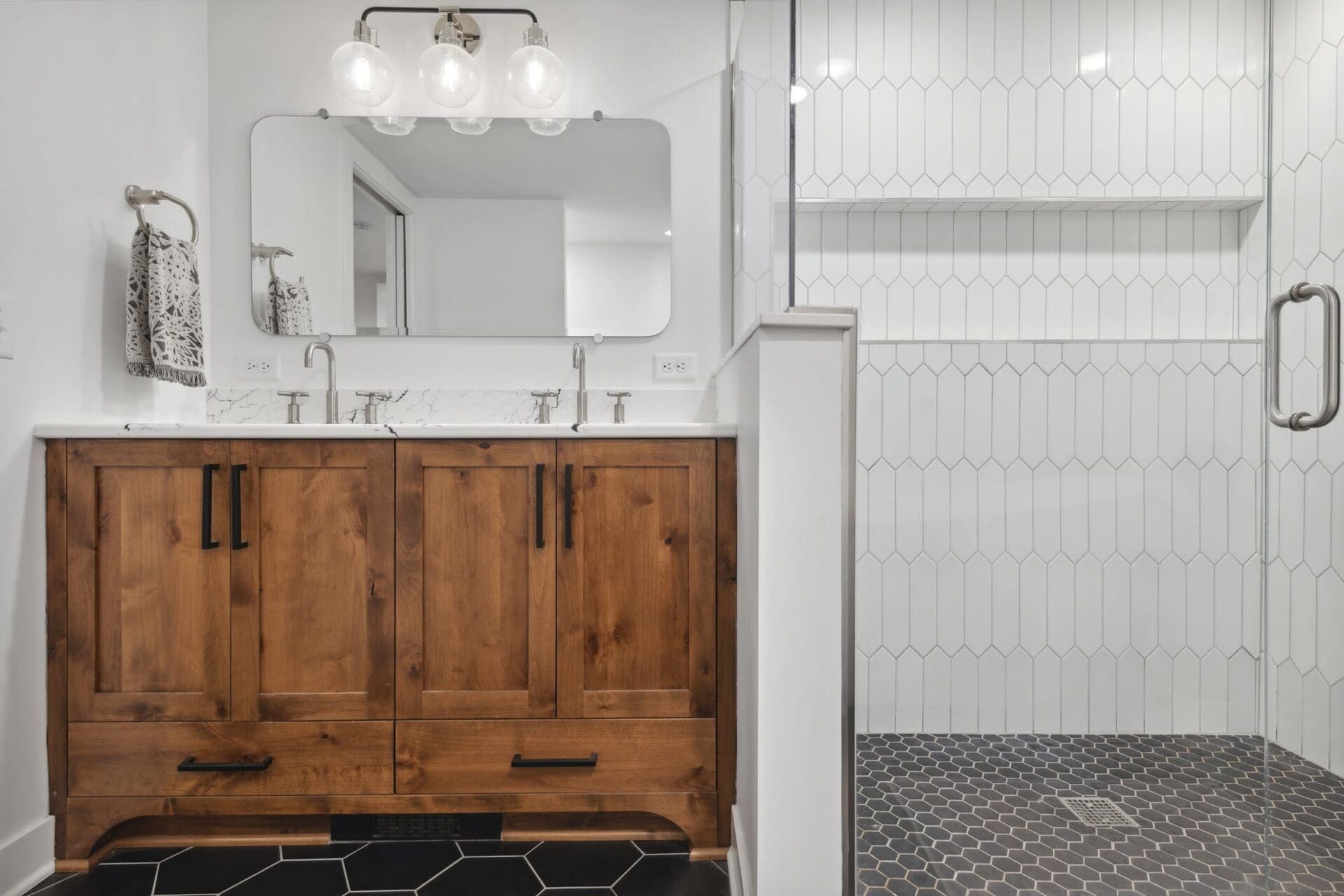 A bathroom with a custom wood double vanity, matte black hardware, white walls, and a walk-in shower featuring white elongated hexagon tile and black hex floor tile.