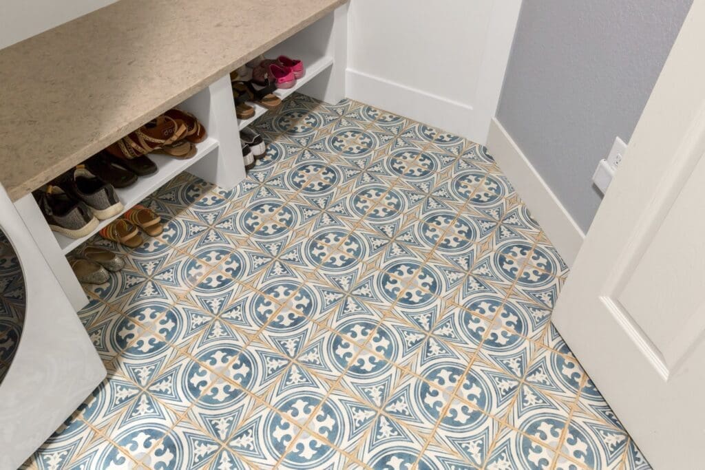 A mudroom entry with white built-in shoe storage and a tan countertop, accented by vibrant patterned floor tile in blue, cream, and gold tones.