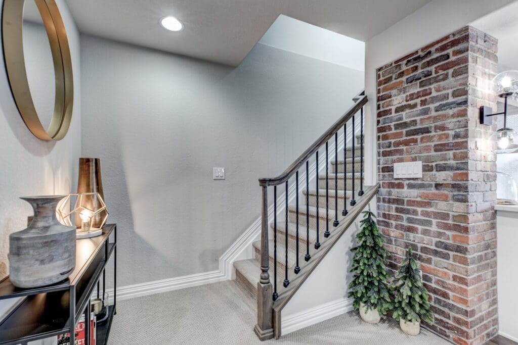 A staircase design featuring neutral carpeting, black iron spindles, and a classic wood handrail. The adjacent wall is finished with exposed red brick for added texture.