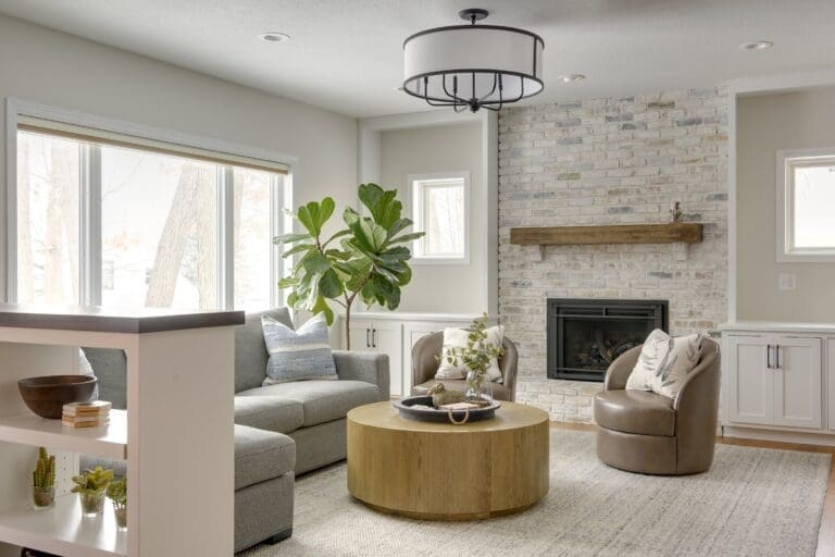 A cozy living room with a light-washed brick fireplace, wooden mantel, built-in cabinets, a gray sectional, a round wooden coffee table, and a fiddle leaf fig tree by the window.